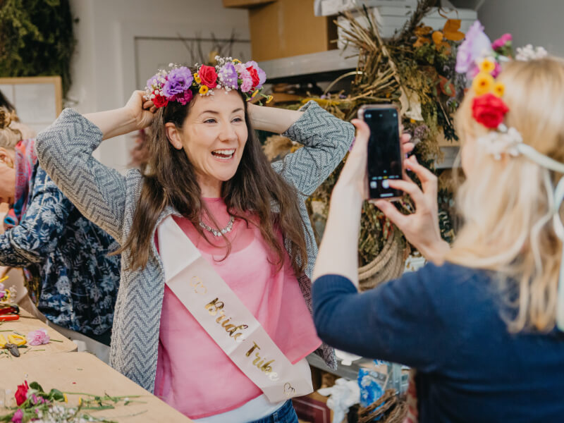 A smiling woman in a bridal sash tries on a flower crown